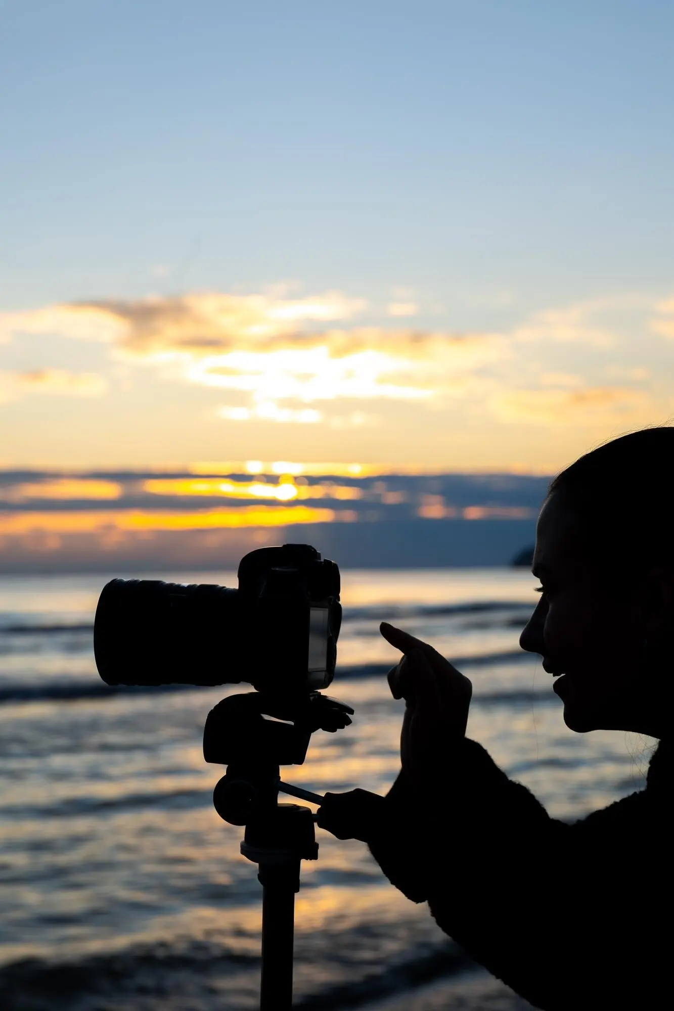 Ein Mädchen stellt bei Sonnenuntergang beim Strand-Shooting am Meer eine Kamera auf einem Stativ ein.